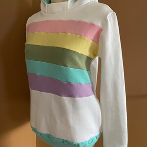 May include: White hoodie with a rainbow stripe pattern in pastel pink, yellow, green, blue, and purple. The hoodie has a light blue hood lining.