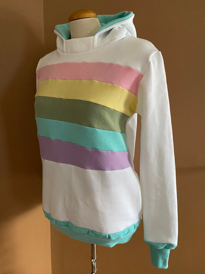 May include: White hoodie with a rainbow stripe pattern in pastel pink, yellow, green, blue, and purple. The hoodie has a light blue hood lining.