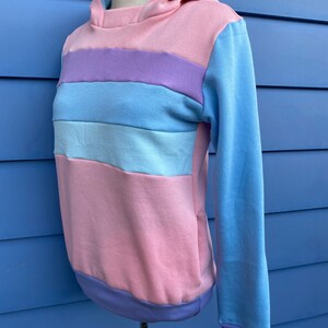 May include: A pastel striped hoodie with a light blue hood and long sleeves. The hoodie has a pink, light blue, and purple striped pattern.
