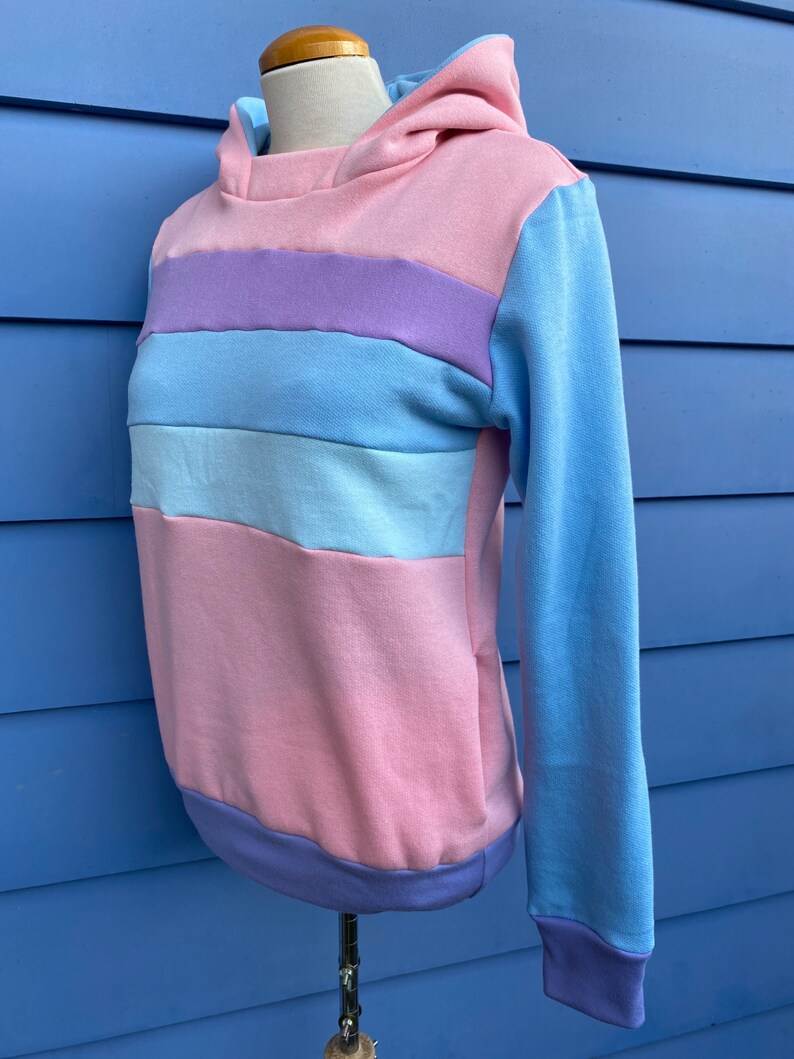 May include: A pastel striped hoodie with a light blue hood and long sleeves. The hoodie has a pink, light blue, and purple striped pattern.