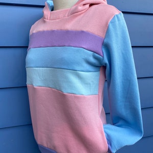 May include: A striped hoodie with a pink, light blue, and purple color scheme. The hoodie has a hood and long sleeves.