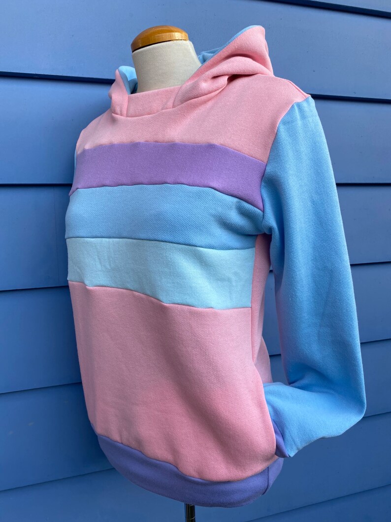 May include: A striped hoodie with a pink, light blue, and purple color scheme. The hoodie has a hood and long sleeves.