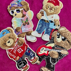 May include: Four embroidered teddy bear patches featuring distinct outfits. One wears a butterfly-print hoodie and sunglasses, another a red cap and glasses, one a red jersey with the text NUMBER 01, and the last a black jacket playing a red guitar.