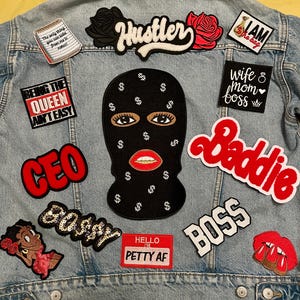 May include: Light blue denim jacket adorned with various patches. Patches include the word "Hustler", a black balaclava with dollar signs, and the word "Baddie". Other patches feature phrases like "CEO" and "Boss".