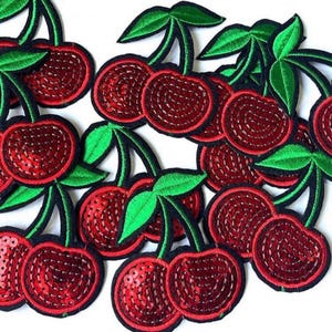 May include: A collection of red sequined cherry patches with green leaves and black stems. The cherries are embroidered with a detailed pattern, creating a textured appearance. These patches are ideal for adding a decorative touch to clothing or accessories.