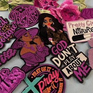 May include: A collection of embroidered patches in shades of purple, black, and brown. The patches feature text like "Bougie AF", "God Don't Play About Me", and illustrations of women. Floral embellishments are also present.