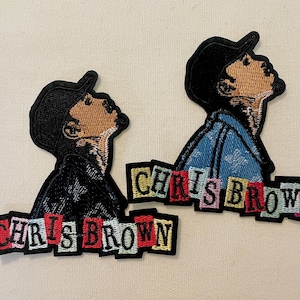 May include: Two embroidered patches featuring a stylized portrait of a person wearing a cap, looking upwards. The patches have the name "CHRIS BROWN" in colorful block letters below the portrait. One patch has a black jacket, the other a blue denim jacket.