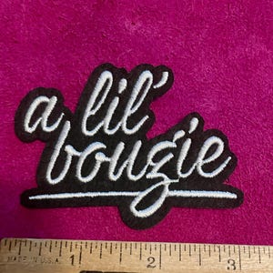 May include: A black embroidered patch with white cursive text that reads a lil bougie. The patch is positioned above a wooden ruler showing a width of approximately 3.5 inches. The embroidery features a clean script font with a solid black background.