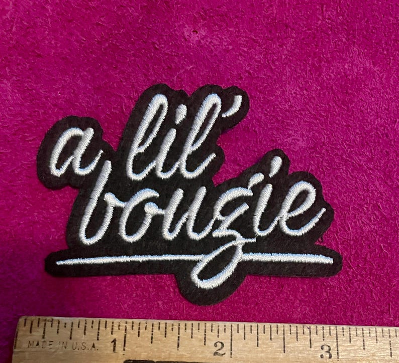 May include: A black embroidered patch with white cursive text that reads a lil bougie. The patch is positioned above a wooden ruler showing a width of approximately 3.5 inches. The embroidery features a clean script font with a solid black background.