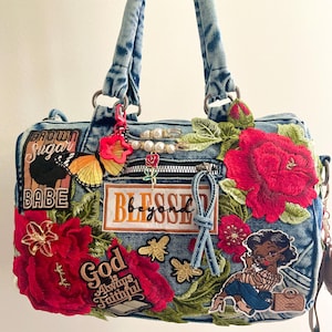May include: A blue denim handbag decorated with various patches including red roses, a butterfly, bees, and a cartoon figure. Text patches read Brown Sugar Babe, Blessed beyond, and God is Always Faithful. The bag features pearl-accented safety pins.