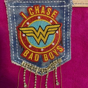 May include: A blue denim pocket patch featuring a red and light blue circular emblem with a yellow Wonder Woman logo, the text "I CHASE BAD BOYS," and white stars. A wooden ruler shows the patch is approximately 4.5 inches wide with crystal fringe.