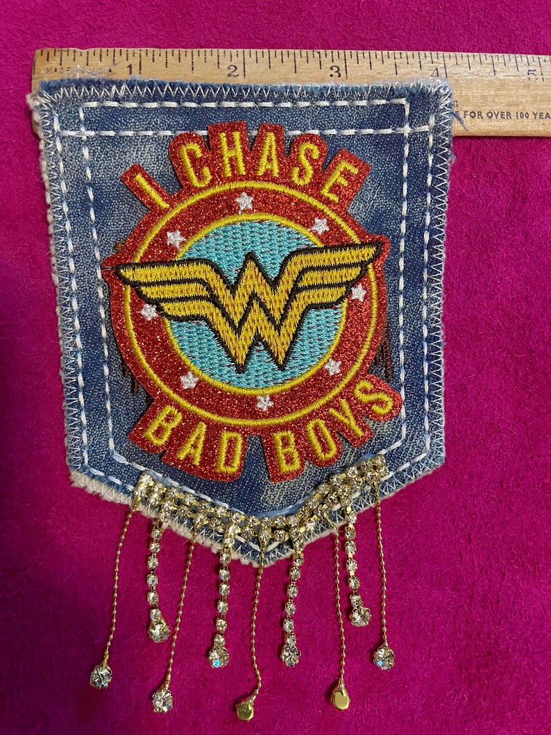 May include: A blue denim pocket patch featuring a red and light blue circular emblem with a yellow Wonder Woman logo, the text "I CHASE BAD BOYS," and white stars. A wooden ruler shows the patch is approximately 4.5 inches wide with crystal fringe.