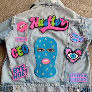 May include: Light blue denim jacket with multiple patches. Patches include the word "Hustler" in pink and green, a blue ski mask with dollar signs, and the words "CEO" and "Bye Hoe!" in pink and green.