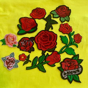 May include: A collection of embroidered rose patches in various sizes and shades of red and pink. The patches feature detailed green leaves and stems, with one rose design including a grey banner with illegible black text at the bottom.