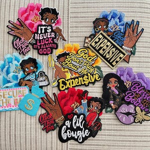 May include: Embroidered patches featuring cartoon characters, floral designs, and text. The patches include phrases like "It's Never Luck, It's Always God" and "Expensive." Colors include pink, blue, yellow, red, and purple. The patches have a variety of designs.