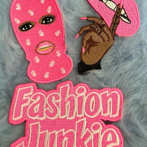 May include: Three pink and white embroidered iron-on patches. One patch features a pink ski mask with white dollar signs and a woman's face. Another patch features a pink hand with a pink fingernail and a pink lip. The third patch features the words "Fashion Junkie" in white letters.