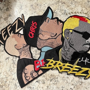 May include: Embroidered patches featuring stylized portraits of a person with the word "Breezy" in gold script. One patch has the name "Chris" in red. The patches have black, gold, and tan colors.