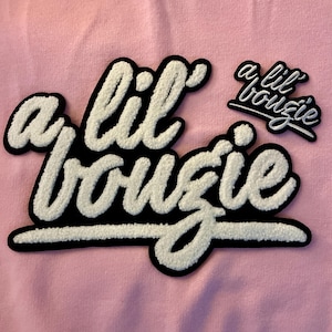 May include: Close-up of a pink fabric with two embroidered patches. The larger patch features the phrase "a lil bougie" in white, textured lettering with a black border. A smaller patch repeats the phrase.