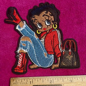 May include: An embroidered patch featuring a cartoon character with black hair, wearing a red jacket, blue distressed jeans, and red high-heeled boots, sitting next to a brown patterned handbag, measured against a ruler showing 3.5 inches.