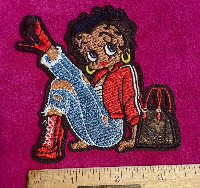 May include: An embroidered patch featuring a cartoon character with black hair, wearing a red jacket, blue distressed jeans, and red high-heeled boots, sitting next to a brown patterned handbag, measured against a ruler showing 3.5 inches.