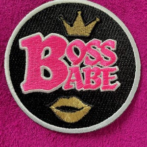 May include: A circular embroidered patch with a black background and a white border. The text BOSS BABE is stitched in bright pink. Above the text is a gold crown, and below the text is a gold pair of lips. The patch is on a textured pink surface.