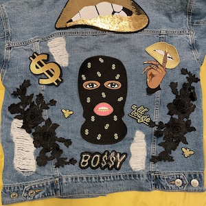 May include: A light blue denim jacket with various patches and embellishments. Features a gold sequined lips patch, a black balaclava patch with dollar signs, and the word "BOSS$Y". Includes black floral and bee accents.