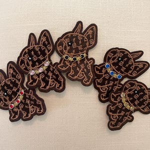 May include: Embroidered patches featuring a brown dog design with a repeating pattern. Each patch has a decorative collar with colorful gems. The patches are arranged on a neutral background.