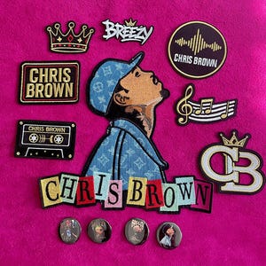 May include: A collection of embroidered patches and circular pins featuring Chris Brown imagery. The set includes a large portrait patch, a crown, a cassette tape, a music note, a logo, and text patches reading BREEZY and CHRIS BROWN.