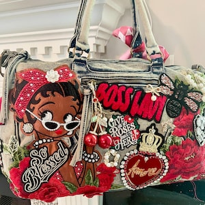 May include: A light blue acid-wash denim handbag featuring a large embroidered patch of a cartoon character with a red headband, the text "So Blessed," "BOSS LADY," and "GIVE ME MY ROSES WHILE I'M HERE," plus red floral, cherry, crown, and heart-shaped patches.
