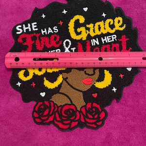 May include: A tufted textile wall hanging featuring a profile of a person with dark skin, red lips, and yellow hoop earrings, surrounded by red roses. The black background includes the text 'SHE HAS FIRE AND GRACE IN HER HEART' and small white stars. A pink ruler shows a width of approximately 30 cm.