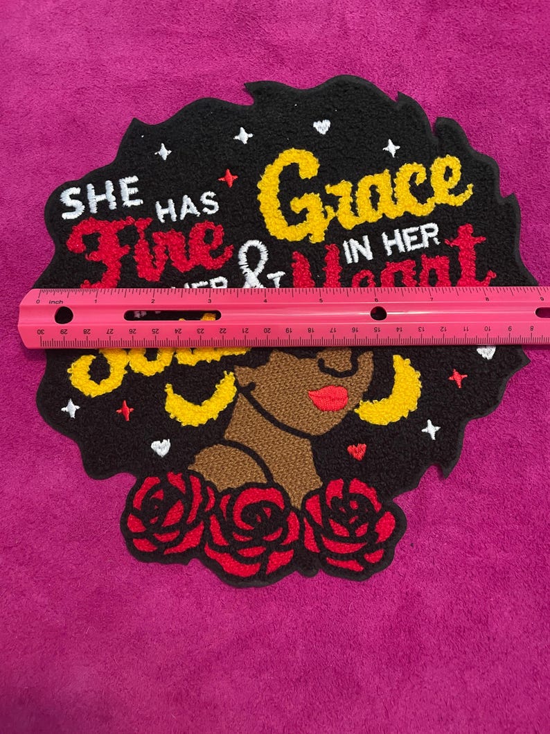 May include: A tufted textile wall hanging featuring a profile of a person with dark skin, red lips, and yellow hoop earrings, surrounded by red roses. The black background includes the text 'SHE HAS FIRE AND GRACE IN HER HEART' and small white stars. A pink ruler shows a width of approximately 30 cm.