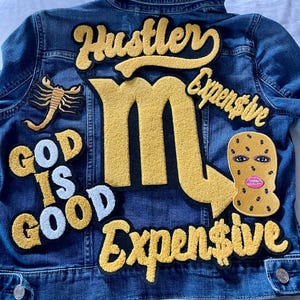 May include: Blue denim jacket with yellow embroidered patches. Patches include the words "Hustler" and "Expensive", a scorpion, a ski mask, and the phrase "God is Good". The large letter "M" is also featured.