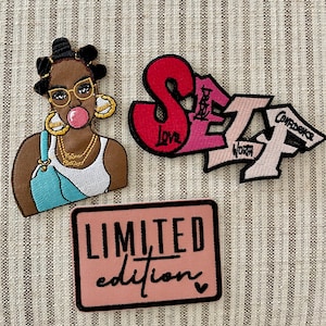 May include: Three embroidered patches: a pink, red, and white patch with the word "SELF" in graffiti style with the words "Love", "Worth", and "Confidence" written on the patches, a pink patch with the words "LIMITED edition" in black, and a cartoon illustration of a Black woman with pink bubblegum, gold hoop earrings, and a blue and white tank top.