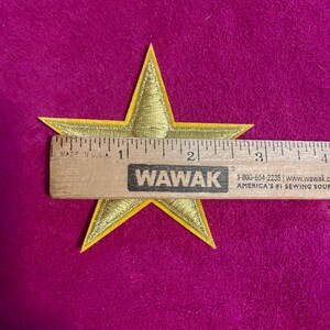 May include: A gold star-shaped embroidered patch with a yellow border. A wooden ruler is placed across the center of the star, showing a width of approximately 3.5 inches from the left point to the right point of the star's horizontal span.