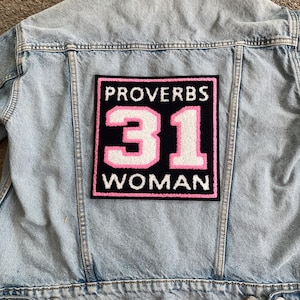 May include: Light blue denim jacket with a large patch on the back. The patch is black with pink and white text that reads "PROVERBS 31 WOMAN". The jacket has a classic design with a button closure and pockets.