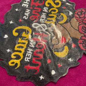 May include: A black, scalloped-edge patch featuring a graphic of a person with red roses in their hair. The text reads, "She has fire in her soul and grace in her heart," printed in yellow and red script. Small white stars are scattered throughout.