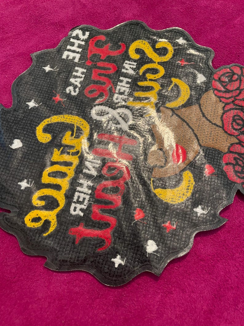 May include: A black, scalloped-edge patch featuring a graphic of a person with red roses in their hair. The text reads, "She has fire in her soul and grace in her heart," printed in yellow and red script. Small white stars are scattered throughout.