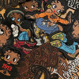 May include: A collection of embroidered patches featuring cartoon characters in various poses. The patches showcase a range of colors, including brown, orange, blue, and black. Some patches include text like "Diva" and "1913".