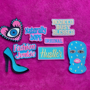 May include: A collection of embroidered patches including a heart with an eye, a high-heeled shoe, and rectangular patches with the text "Naturally Dope," "Booked Busy Blessed," "Original," "Fashion Junkie," "Hustler," and a blue ski mask face.