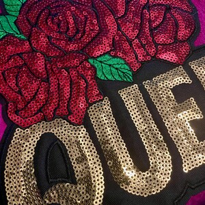 May include: A decorative patch featuring a large red sequined rose with green sequined leaves positioned above the gold sequined text QUE. The design is set against a black fabric background, which is layered over a vibrant magenta textile surface.