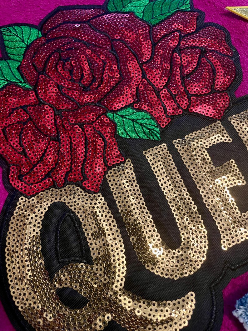May include: A decorative patch featuring a large red sequined rose with green sequined leaves positioned above the gold sequined text QUE. The design is set against a black fabric background, which is layered over a vibrant magenta textile surface.