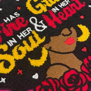 May include: A black tufted rug featuring the embroidered text "HAS GRACE IN HER SOUL & FIRE IN HER HEART" in yellow, red, and white. The design includes a brown silhouette of a person, red roses, yellow crescent moons, and small white stars.