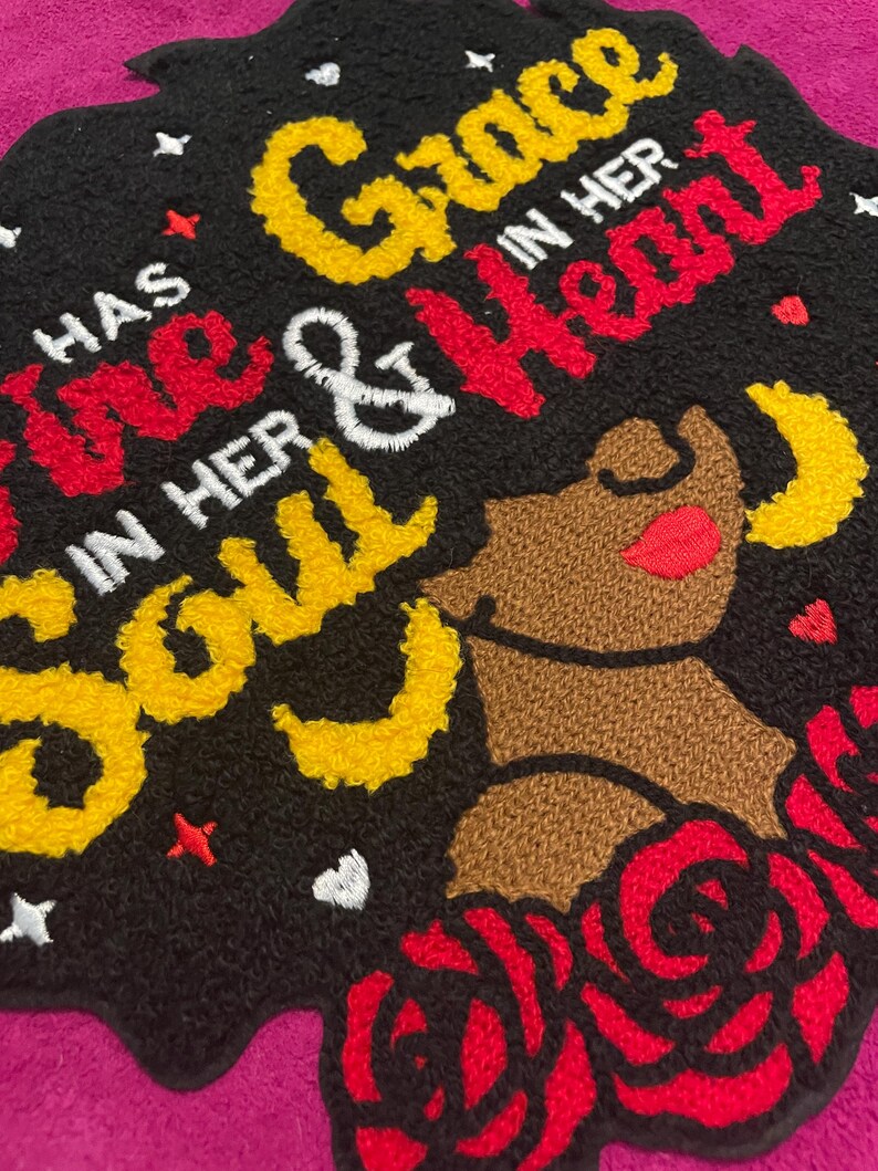 May include: A black tufted rug featuring the embroidered text "HAS GRACE IN HER SOUL & FIRE IN HER HEART" in yellow, red, and white. The design includes a brown silhouette of a person, red roses, yellow crescent moons, and small white stars.