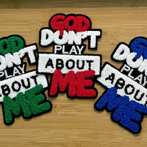 May include: Embroidered patches with the phrase "GOD DON'T PLAY ABOUT ME" in white, red, green, and blue. The patches have a black border and are displayed on a wooden surface. The patches are a decorative item.