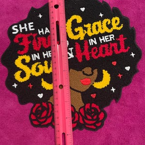 May include: A tufted wall hanging featuring a black silhouette of a person with large hair, red roses, and the text "SHE HAS FIRE IN HER SOUL AND GRACE IN HER HEART". A pink ruler is placed vertically over the piece, measuring approximately 30 cm.