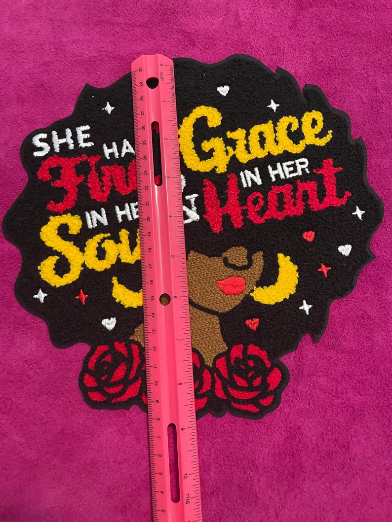 May include: A tufted wall hanging featuring a black silhouette of a person with large hair, red roses, and the text "SHE HAS FIRE IN HER SOUL AND GRACE IN HER HEART". A pink ruler is placed vertically over the piece, measuring approximately 30 cm.