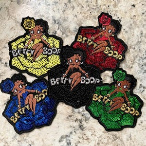 May include: Five embroidered patches featuring Betty Boop in various colors: yellow, red, black, blue, and green. Each patch has a sequined flower design and the words "Betty Boop" in gold lettering.
