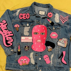 May include: A distressed denim jacket adorned with various patches. Patches include the word "Hustler", "CEO", a pink ski mask, lipstick, lips, a stack of cash, and a high heel. The jacket has a button-up front and two chest pockets.