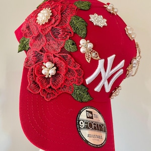 May include: A red baseball cap with embroidered red roses and green leaves. The cap features white accents, pearl embellishments, and a white "NY" logo. The cap is adjustable and has a "9FORTY" label.