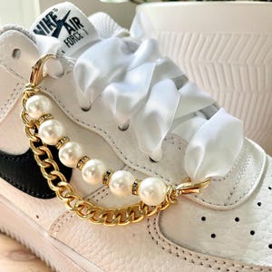 May include: White leather sneaker with a gold chain and pearl shoe accessory. The accessory features large faux pearls and small rhinestone accents. The sneaker has white laces and the words "NIKE AIR FORCE" printed on the tongue.